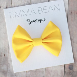 May include: A bright yellow fabric bow tie. The bow tie is on a white card with the text "Emma Bean Boutique".
