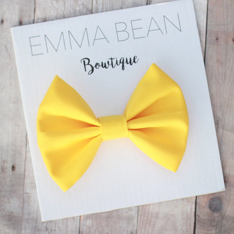 May include: A bright yellow fabric bow tie. The bow tie is on a white card with the text "Emma Bean Boutique".