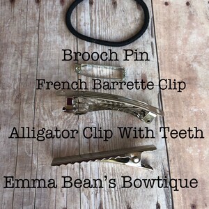 May include: A close-up of four different hair accessories on a wooden surface. The accessories include a ponytail band, a brooch pin, a French barrette clip, and an alligator clip with teeth. The text "Emma Bean's Bowtique" is visible at the bottom of the image.