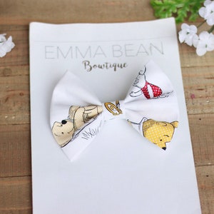 May include: White fabric bow with a Winnie the Pooh print featuring Winnie the Pooh, Piglet, and Tigger. The bow is tied and ready to be worn. The text "Emma Bean Boutique" is printed on the white background.