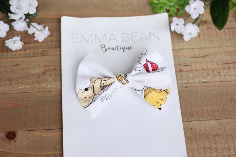 May include: White fabric bow with a Winnie the Pooh print featuring Winnie the Pooh, Piglet, and Tigger. The bow is tied and ready to be worn. The text "Emma Bean Boutique" is printed on the white background.