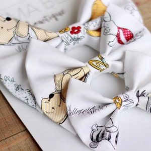 May include: Two white fabric hair bows with a Winnie the Pooh and friends print. The bows feature yellow, brown, and red accents.