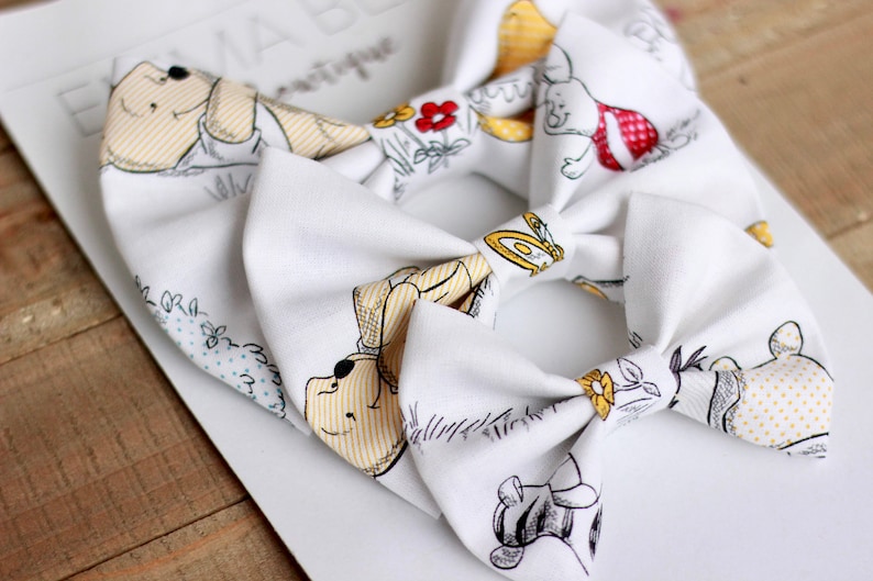 May include: Two white fabric hair bows with a Winnie the Pooh and friends print. The bows feature yellow, brown, and red accents.