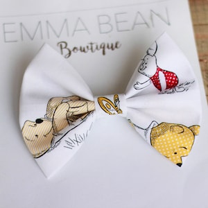 May include: White fabric bow with a Winnie the Pooh print featuring Winnie the Pooh, Piglet, and Tigger. The bow is tied with a white ribbon.