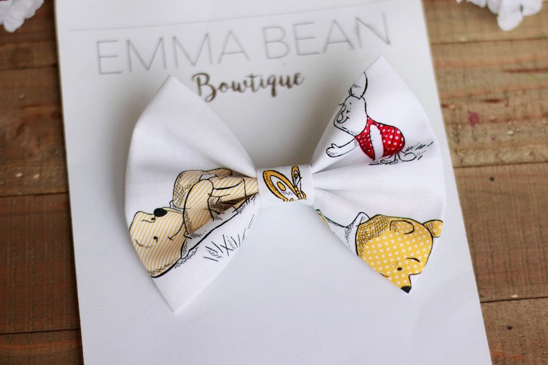 May include: White fabric bow with a Winnie the Pooh print featuring Winnie the Pooh, Piglet, and Tigger. The bow is tied with a white ribbon.