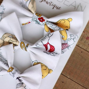 May include: Two white fabric hair bows with a Winnie the Pooh and Piglet print. The bows feature yellow, red, and black accents.