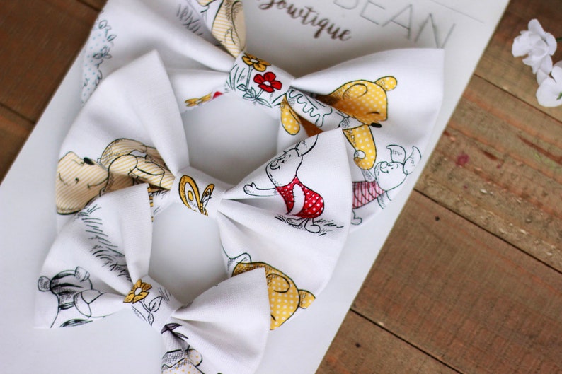 May include: Two white fabric hair bows with a Winnie the Pooh and Piglet print. The bows feature yellow, red, and black accents.
