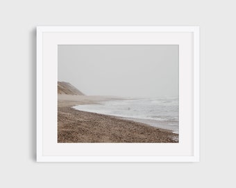May include: A photograph of a beach with a sandy shore and a grey sky. The waves are crashing on the shore and there is a small hill in the distance.