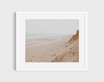 May include: A photograph of a sandy beach with a large sand dune in the foreground. The beach is empty and the water is calm. The sky is overcast and the sand is a light brown color.