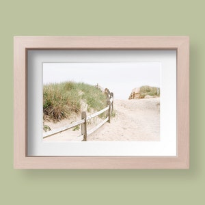 May include: A wooden fence leads to a sandy path through tall grass dunes. The path leads to a beach with a cloudy sky.