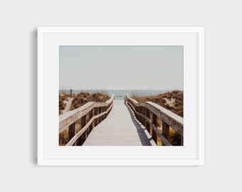 May include: A wooden walkway with railings leads to the beach. The walkway is surrounded by sand dunes and tall grass. The ocean is visible in the distance.
