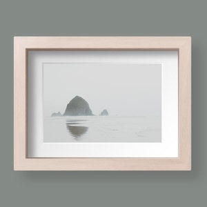 May include: A framed photograph of a rocky island in the ocean. The island is covered in fog and the water is calm. The frame is made of light wood.