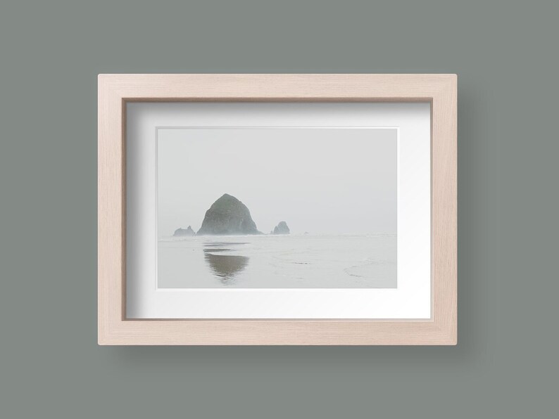 May include: A framed photograph of a rocky island in the ocean. The island is covered in fog and the water is calm. The frame is made of light wood.