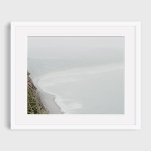 May include: A misty, aerial view of a coastline with a rocky cliff and a sandy beach. The ocean is a light blue and the sky is a hazy white.