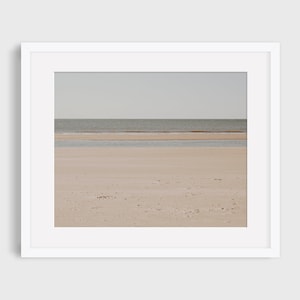 Puede incluir: Una fotografía de una playa de arena con un mar tranquilo en el fondo. La arena es de color marrón claro y el agua es de color azul claro. El cielo es de color gris claro.