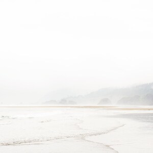 May include: A misty beach scene with a coastline in the distance. The water is calm and the sand is light brown. The sky is a pale blue and the air is hazy.