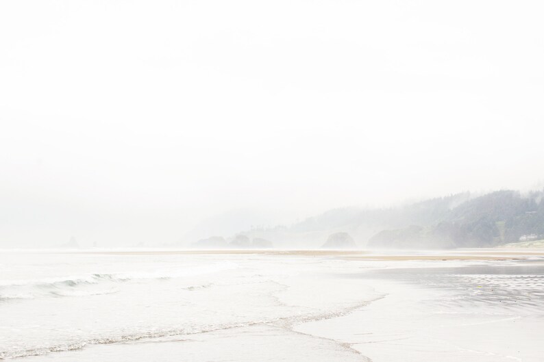 May include: A misty beach scene with a coastline in the distance. The water is calm and the sand is light brown. The sky is a pale blue and the air is hazy.