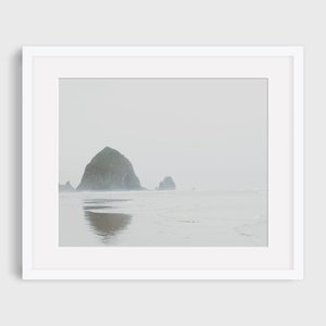 May include: A misty seascape with a large rock formation in the distance, reflecting in the water. The sky is a pale gray, and the water is a light blue.