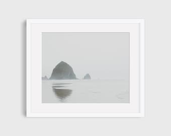 May include: A misty seascape with a large rock formation in the distance, reflecting in the water. The sky is a pale gray, and the water is a light blue.