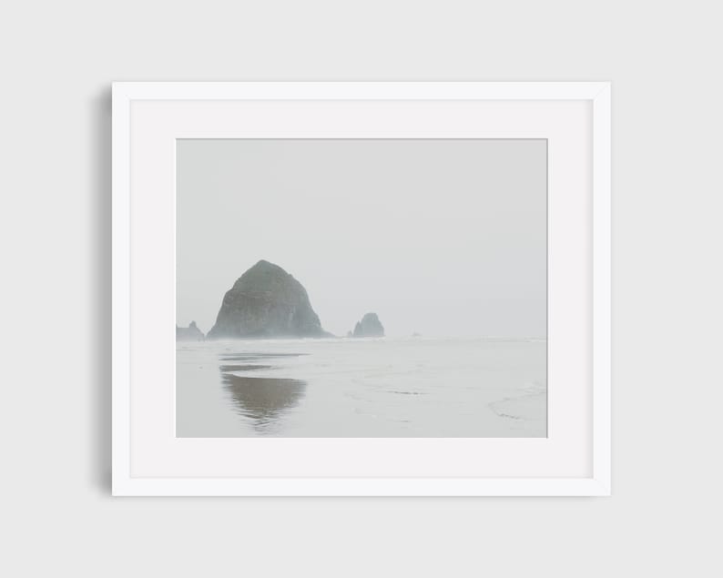 May include: A misty seascape with a large rock formation in the distance, reflecting in the water. The sky is a pale gray, and the water is a light blue.