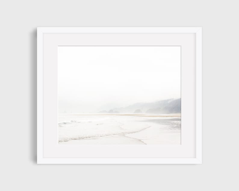 May include: A photograph of a misty beach scene with waves lapping at the shore. The water is a light blue and the sand is a pale yellow. The background is a hazy, light blue sky with a line of trees in the distance.