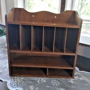 May include: A brown wooden wall-mounted organizer featuring a top section with five vertical dividers and a bottom section divided into two horizontal compartments. The unit has a scalloped top edge and two pre-drilled holes for mounting purposes.