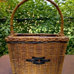 May include: A rectangular wicker basket with a curved handle. The basket is woven with light brown and black wicker, featuring a geometric design on the front. The handle is made of a thicker, lighter-colored wood. The basket is designed for carrying items.
