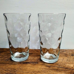 May include: Two clear glass tumblers featuring a textured exterior surface with a repeating pattern of circular indentations. Each drinking glass has a slightly tapered cylindrical shape with a thick, smooth base and a wide, open rim.