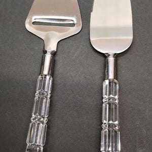 May include: Two serving utensils: a cheese slicer and a cake server. Both have shiny, silver-colored blades and clear, faceted handles. The cheese slicer has a rectangular blade with a slot, while the cake server has a triangular blade with a serrated edge.