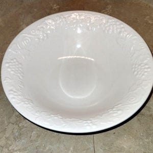 May include: A white, round serving bowl with a decorative embossed grape and vine pattern around the rim. The bowl has a smooth, glossy finish and a shallow depth, suitable for serving salads or side dishes.