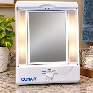 May include: A white Conair lighted vanity mirror with a rectangular frame and vertical side lights. The base features a circular control knob with settings labeled Day, Evening, Home, Office, and Bright, allowing for adjustable lighting.