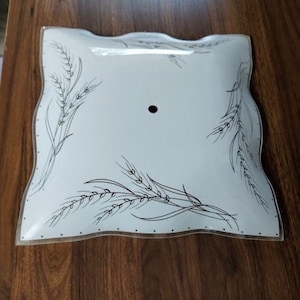 May include: A square, white glass light fixture cover with wavy edges and a central circular hole. The surface features a black line art illustration of wheat stalks arranged in the corners, complemented by a delicate dotted border along the edges.
