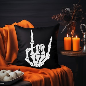 May include: Black throw pillow with a white skeleton hand making a rock and roll hand gesture. The pillow is on an orange throw blanket.