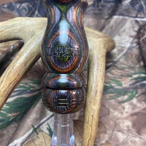 May include: A wooden duck call with a green, black, and brown camouflage pattern. The call has a clear plastic insert and the text "TACHETT GAME CALLS" is engraved on the side.