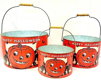 May include: Set of three vintage-style Halloween buckets. The red metal buckets feature a smiling jack-o'-lantern design with the words "Happy Halloween." Each bucket has a wire handle with a wooden grip.