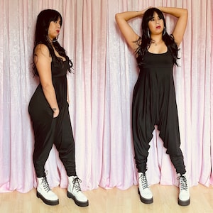 May include: A woman wearing a black jumpsuit with a wide leg and a low cut neckline. She is wearing white platform boots and has her hair pulled back. The background is a pink curtain.