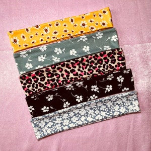 May include: Five rectangular fabric headbands arranged in a stack. The top headband is yellow with white daisy flowers. The second is sage green with white flowers. The third features a pink and black leopard print. The fourth is black with white flowers. The bottom is light gray with white flowers.