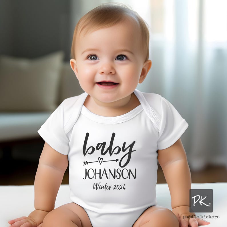 a baby sitting on a bed wearing a white announcement onesie that says baby JOHANSON Winter 2024