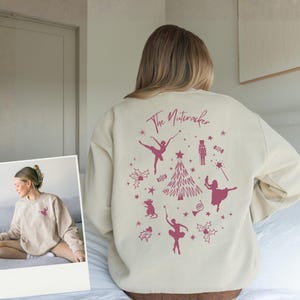 Op de afbeelding: Crèmekleurige sweatshirt met een roze afbeelding van balletdansers, een kerstboom en de tekst "The Nutcracker". Het ontwerp bevat sterren, een muis en een notenkrakerfiguur. De sweatshirt heeft een relaxte pasvorm.