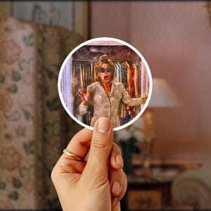 May include: A circular image of a woman wearing sunglasses and a tan suit, with her mouth open in surprise, is held by a hand with a gold ring. The background shows a closet with clothes and a room with a lamp and sofa.