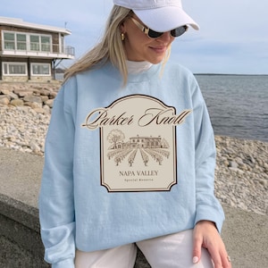 Può includere: Una felpa girocollo azzurro chiaro con una stampa grafica centrale bordata in marrone scuro. Il design include la scritta Parker Knoll sopra un dettagliato disegno al tratto di un vigneto e un edificio, con la scritta NAPA VALLEY Special Reserve.