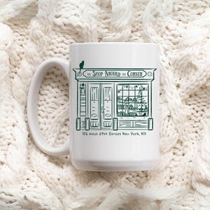 Op de afbeelding: Witte keramische mok met een groene illustratie van een winkelgevel, de tekst "The Shop Around the Corner" en het adres "106 West 69th Street New York, NY". Een zwarte kat zit bovenop het bord.