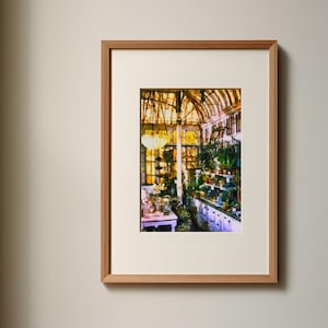 Puede incluir: Una impresión enmarcada que muestra el interior de un invernadero soleado con techo de cristal, numerosas plantas en macetas sobre estantes y una mesa blanca. La obra está en un marco de madera clara con un paspartú ancho de color blanco roto.