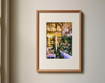 Op de afbeelding: Een ingelijste print met het interieur van een zonnige kas met een glazen dak, talloze potplanten op planken en een witte tafel. Het kunstwerk is geplaatst in een lichtgekleurde houten lijst met een brede gebroken witte passe-partout.