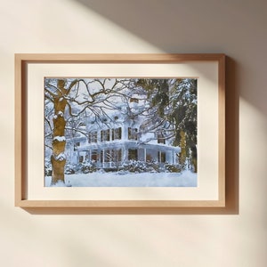 May include: A framed watercolour painting depicting a white house with snow-covered trees in the foreground. The house features a porch with a railing and a roof with a dormer window. The trees are bare and the snow is white.