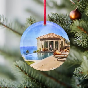 Puede incluir: Un adorno redondo de vidrio con una pintura de acuarela de una casa blanca con una piscina y un cielo azul. La casa tiene un techo de tejas rojas y un patio con sillas.