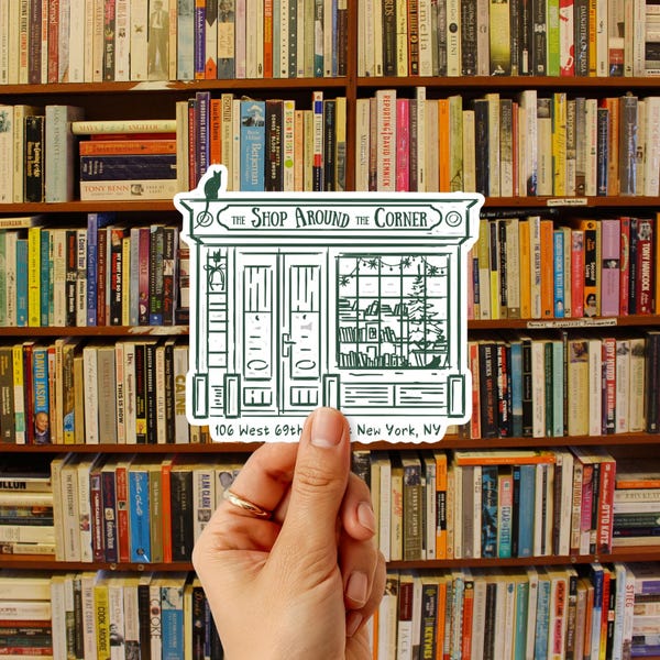 May include: A die-cut sticker featuring a dark green line drawing of a storefront labelled The Shop Around the Corner, 106 West 69th St, New York, NY. The illustration depicts a shop facade with a cat on the roof and a window display of books.
