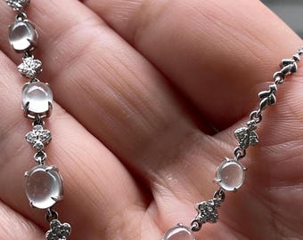 May include: A silver-toned necklace featuring a series of oval-shaped, translucent white cabochon stones alternating with small, four-petaled floral links encrusted with tiny clear stones. The chain is constructed with delicate metal links.