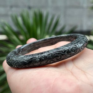 May include: A dark gray, carved stone bracelet with intricate floral and swirl patterns. The bracelet is circular and appears to be made of a solid, polished material. The design is consistent around the entire band.