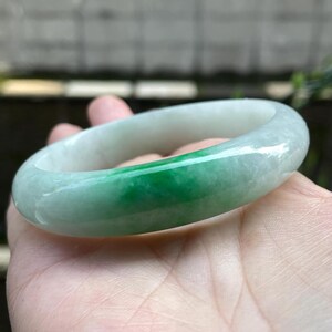 May include: A jade bangle bracelet with a smooth, polished finish. The bracelet displays a colour gradient, shifting from a pale, near-white shade to a vivid, deep green. The jade is a solid, opaque material.
